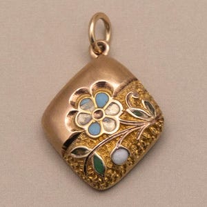 May include: A gold-coloured pendant in a diamond shape. It features a floral design with blue, white, and green accents. The flower is in the centre, with leaves and a small white bead. A small loop is at the top for a chain.