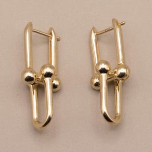 Peut inclure: Une paire de boucles d'oreilles créoles en or poli au design en forme de maillon en U. Chaque boucle se compose d'une boucle métallique lisse et allongée reliée à une barre horizontale centrale avec deux petits accents sphériques sur les côtés.