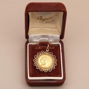 May include: A gold pendant with a Native American profile in a decorative filigree frame. The pendant is in an open, brown velvet jewellery box with the text "J. Byrons 14 KT. GOLD" on the inside.