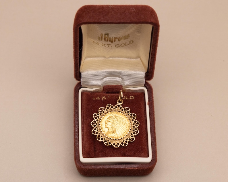 May include: A gold pendant with a Native American profile in a decorative filigree frame. The pendant is in an open, brown velvet jewellery box with the text "J. Byrons 14 KT. GOLD" on the inside.