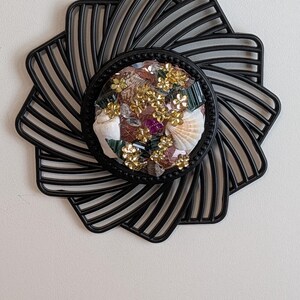 Op de afbeelding: Een decoratieve zwarte metalen onderzetter met een cirkelvormig middenstuk met schelpen, goudkleurige bloemaccenten en kleurrijke edelstenen. De onderzetter heeft een uniek, gelaagd ontwerp dat lijkt op een gestileerde bloem.