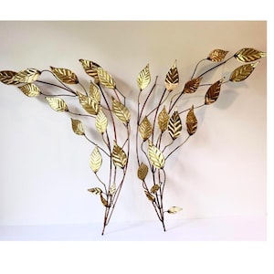 May include: A pair of wall-mounted metal sculptures featuring two branching stems with numerous polished gold-toned leaves. The branches extend outward in a symmetrical arrangement, showcasing detailed veining on each individual leaf surface.