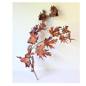 May include: A decorative wall sculpture shaped like a branch with multiple jagged, maple-like leaves. The metal leaves feature a gradient of warm colors, including copper, orange, red, and dark brown tones, with a metallic, reflective finish.