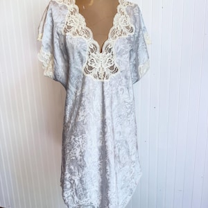 May include: A light gray nightgown featuring a subtle tonal damask pattern, short sleeves, and a deep V-neckline trimmed with intricate cream-colored floral lace. The garment has a relaxed, knee-length silhouette with matching lace trim on the sleeves.