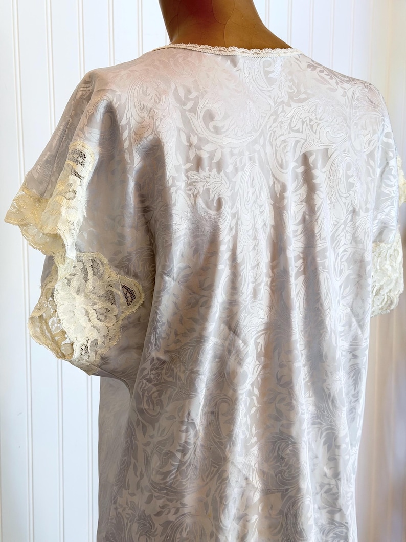 May include: A light gray satin nightgown featuring a tonal paisley jacquard pattern throughout. The garment has short sleeves finished with cream-colored lace trim and a matching lace detail along the neckline, providing a soft texture.