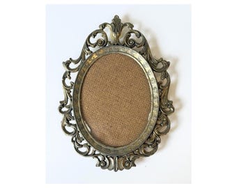 May include: An oval-shaped picture frame featuring an ornate, metallic gold-toned border with intricate scrollwork and leaf-like detailing. The center of the frame contains a textured, tan-colored fabric insert, providing a neutral background.