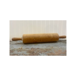 May include: A wooden rolling pin with handles on each end. The rolling pin is made of light brown wood and has a worn, rustic look.
