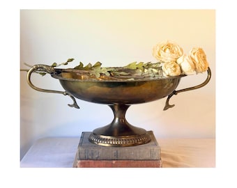 May include: A polished brass pedestal bowl featuring two ornate handles shaped like dolphin tails. The bowl has a wide, shallow basin and a circular base with a ribbed edge. Three cream-colored artificial flowers rest inside the bowl.