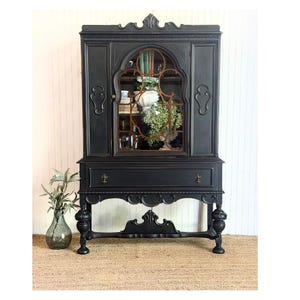 May include: A tall, matte black wooden hutch featuring a single arched glass door with decorative mullions, a wide horizontal drawer with two metal pulls, and four turned legs connected by a carved lower stretcher with a central finial.