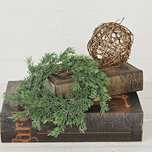 May include: A green artificial cedar wreath resting on a dark brown leather-bound book, accompanied by a spherical brown woven twig orb. The book features decorative gold-toned embossed lettering and lines on its front cover surface.