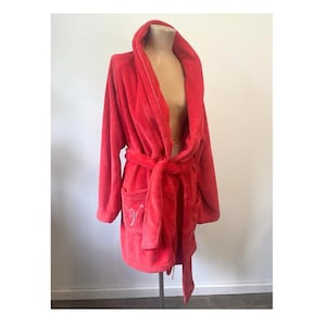 May include: A plush, red robe with a shawl collar and long sleeves. The robe features a matching tie belt and pockets. The robe is displayed on a mannequin. The robe has an embroidered design on the pocket.