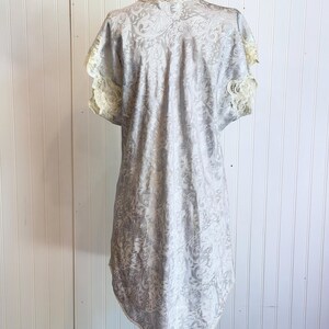 May include: A light gray short-sleeved nightgown featuring a tonal paisley pattern throughout the fabric. The garment includes delicate cream-colored lace trim along the sleeve edges and a scalloped lace neckline, with a rounded hemline.