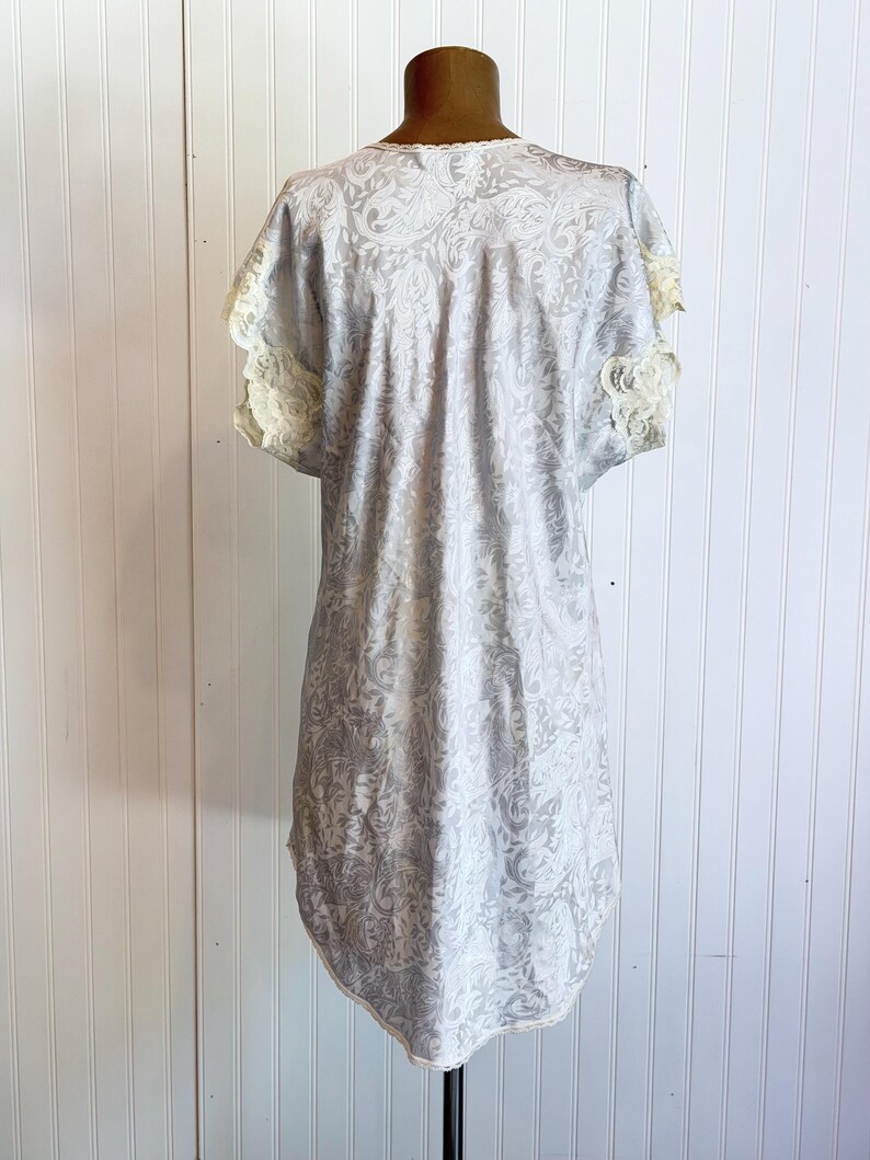 May include: A light gray short-sleeved nightgown featuring a tonal paisley pattern throughout the fabric. The garment includes delicate cream-colored lace trim along the sleeve edges and a scalloped lace neckline, with a rounded hemline.