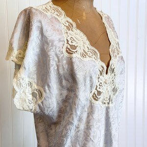 May include: A light blue nightgown featuring a subtle tonal damask pattern throughout the fabric. The garment has short sleeves and a deep V-neckline, both trimmed with intricate cream-colored floral lace detailing along the edges.