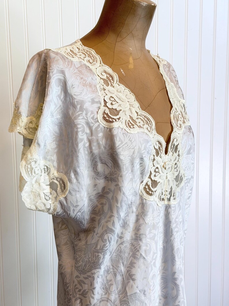 May include: A light blue nightgown featuring a subtle tonal damask pattern throughout the fabric. The garment has short sleeves and a deep V-neckline, both trimmed with intricate cream-colored floral lace detailing along the edges.