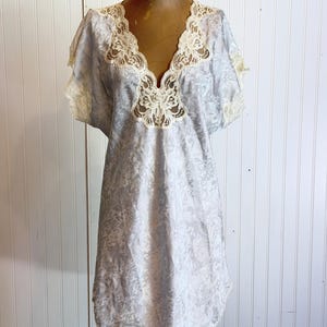 May include: A light blue short-sleeved nightgown featuring a subtle tonal floral jacquard pattern throughout the fabric. The garment has a deep V-neckline trimmed with intricate cream-colored lace, which also accents the sleeve edges.