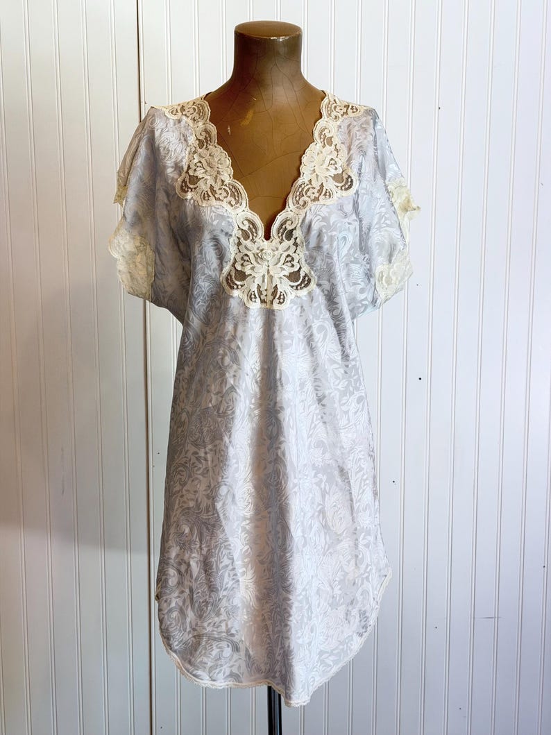 May include: A light blue short-sleeved nightgown featuring a subtle tonal floral jacquard pattern throughout the fabric. The garment has a deep V-neckline trimmed with intricate cream-colored lace, which also accents the sleeve edges.