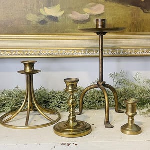 May include: Four gold metal candle holders. The tallest candle holder has a round base and three legs. The other three candle holders have different designs.