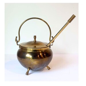 May include: A brass pot with a rounded body, three small legs, and a matching lid with a central knob. It features a curved handle attached to the sides and a long, straight handle extending diagonally from the top rim with a ribbed grip.