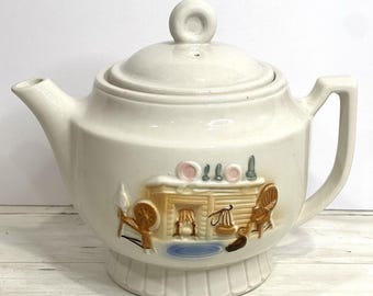 May include: Cream-colored ceramic teapot with a lid and handle. The teapot features a raised relief design of a cabin scene with a fireplace, chairs, and other decorative elements. The teapot has a spout and sits on a pedestal base.