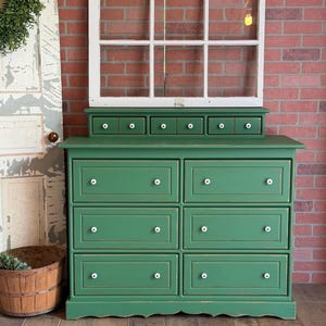 May include: A green dresser with white knobs and six drawers. A small shelf with five drawers sits on top. A white window frame is behind the dresser, set against a red brick wall. A wooden basket and a wreath are on the left.