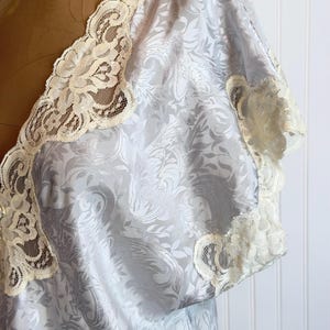 May include: A light blue satin robe featuring a tonal damask floral pattern and cream-colored lace trim along the V-neckline and short sleeves. The garment is displayed on a mannequin, showcasing the intricate lace detailing and the fabric texture.