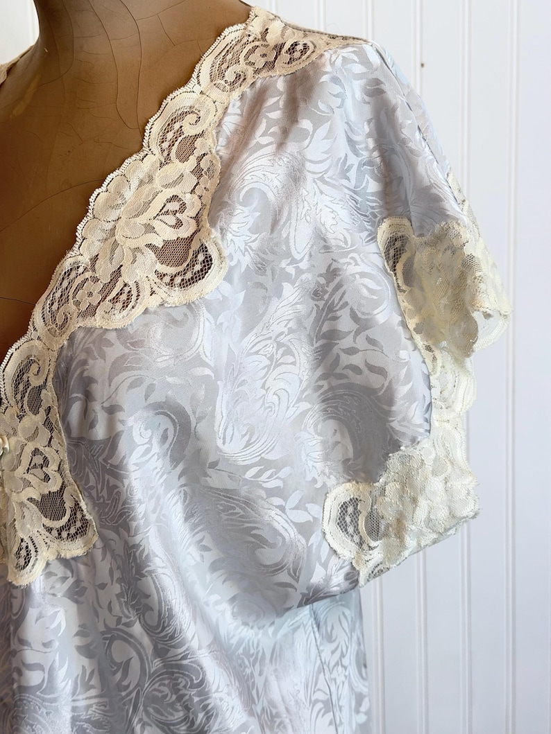 May include: A light blue satin robe featuring a tonal damask floral pattern and cream-colored lace trim along the V-neckline and short sleeves. The garment is displayed on a mannequin, showcasing the intricate lace detailing and the fabric texture.