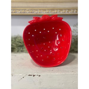 May include: A bright red, strawberry-shaped bowl with small, evenly spaced holes. The bowl has a textured surface and a green leafy top. The bowl is sitting on a white surface.