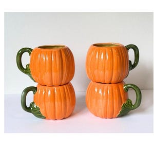 May include: Four ceramic mugs shaped like orange pumpkins with vertical ridges and green handles. Two mugs are stacked on the left side, while two mugs are stacked on the right side. Each mug features a glossy finish and a green leaf detail at the base.
