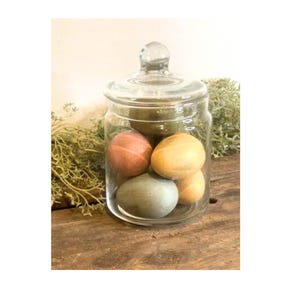 May include: A clear glass jar with a lid, containing four colorful Easter eggs. The eggs are painted in shades of green, yellow, orange, and brown. The jar is sitting on a wooden surface.