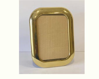 May include: A rectangular, gold-colored picture frame with rounded corners. The frame has a shiny, metallic finish and a cardboard backing. The frame is designed to hold a photograph or artwork and is suitable for tabletop display.