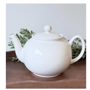 May include: A round, white ceramic teapot with a matching lid featuring a small knob handle. The teapot has a curved spout on the left side and a C-shaped handle on the right side, with a smooth, glossy finish throughout the entire exterior surface.