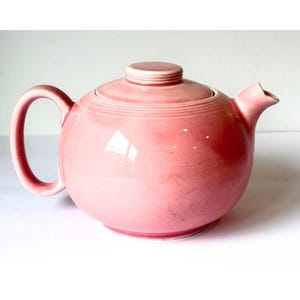 May include: A round, glossy pink teapot featuring a matching lid with concentric ring detailing on the top. The teapot has a curved handle on the left side and a short, pointed spout on the right, with a smooth, solid-colored ceramic finish.