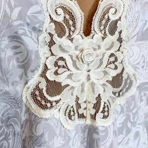May include: A white garment featuring a tonal damask pattern with a prominent cream-colored lace collar. The lace trim extends down the center front and is accented by a small, centered fabric rose detail at the base of the neckline.