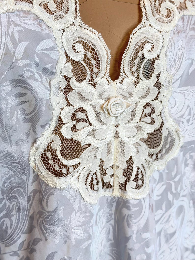 May include: A white garment featuring a tonal damask pattern with a prominent cream-colored lace collar. The lace trim extends down the center front and is accented by a small, centered fabric rose detail at the base of the neckline.