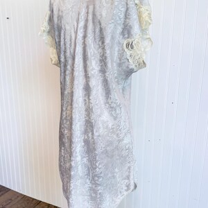 May include: A light gray short-sleeved nightgown featuring a tonal damask floral pattern throughout the fabric. The garment includes cream-colored lace trim along the sleeve edges and the neckline, with a straight hemline and a loose fit.