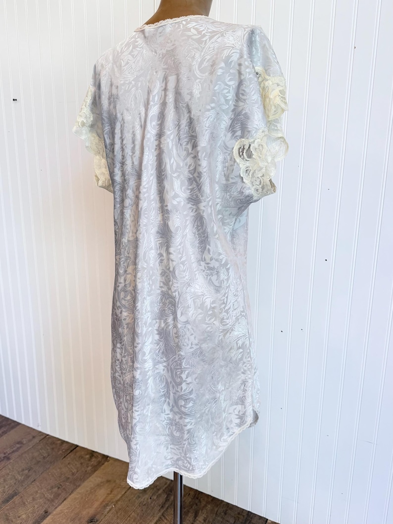 May include: A light gray short-sleeved nightgown featuring a tonal damask floral pattern throughout the fabric. The garment includes cream-colored lace trim along the sleeve edges and the neckline, with a straight hemline and a loose fit.