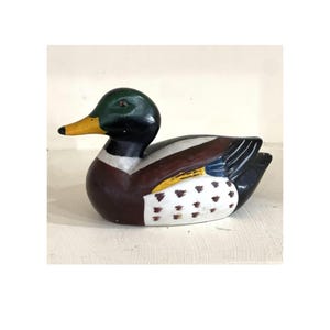 May include: A hand-painted wooden duck figurine. The duck has a green head, a yellow beak, and a brown body with white and black accents. The duck is in a resting position.