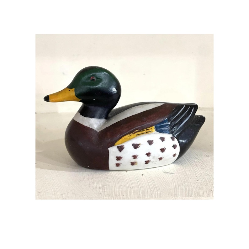May include: A hand-painted wooden duck figurine. The duck has a green head, a yellow beak, and a brown body with white and black accents. The duck is in a resting position.