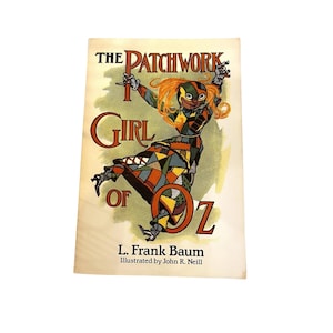 May include: A vintage book cover for "The Patchwork Girl of Oz" by L. Frank Baum, illustrated by John R. Neill. The cover features a colorful illustration of the Patchwork Girl with orange hair and a patchwork outfit. The title is in large, ornate letters.