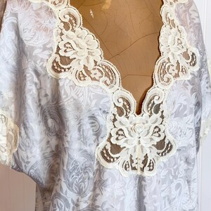 May include: A light gray blouse featuring a tonal damask pattern and a deep V-neckline trimmed with intricate cream-colored lace. The garment includes short sleeves with matching lace trim and a small fabric rose detail at the center chest.