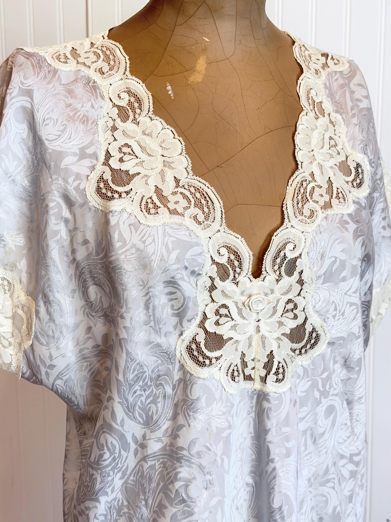 May include: A light gray blouse featuring a tonal damask pattern and a deep V-neckline trimmed with intricate cream-colored lace. The garment includes short sleeves with matching lace trim and a small fabric rose detail at the center chest.