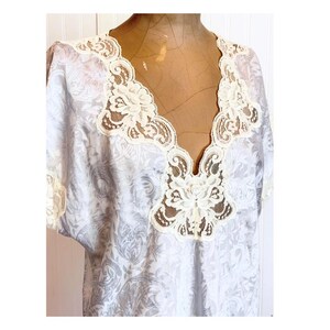 May include: A light gray and white patterned top featuring a deep V-neckline adorned with intricate cream-colored floral lace trim. The garment has short sleeves with matching lace edges and a subtle, repeating damask-style print throughout.