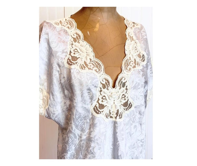 May include: A light gray and white patterned top featuring a deep V-neckline adorned with intricate cream-colored floral lace trim. The garment has short sleeves with matching lace edges and a subtle, repeating damask-style print throughout.