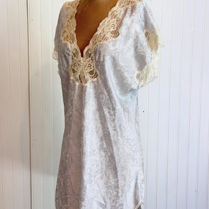 May include: A light blue satin nightgown featuring a subtle tonal floral jacquard pattern, short sleeves, and a deep V-neckline trimmed with cream-colored lace. The garment has a loose, flowing silhouette and reaches mid-thigh length.