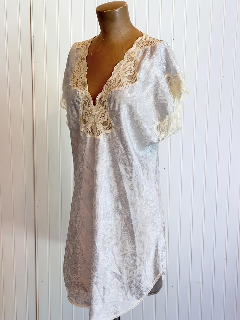 May include: A light blue satin nightgown featuring a subtle tonal floral jacquard pattern, short sleeves, and a deep V-neckline trimmed with cream-colored lace. The garment has a loose, flowing silhouette and reaches mid-thigh length.