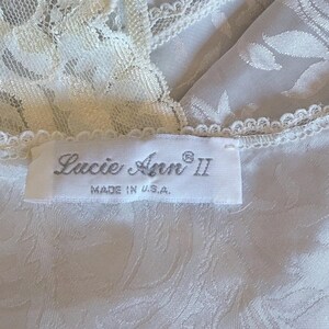May include: A white garment featuring a tonal floral jacquard pattern and cream-colored lace trim along the neckline. A white fabric label is stitched to the garment, displaying the text 'Lucie Ann II' and 'MADE IN U.S.A.' in black lettering.