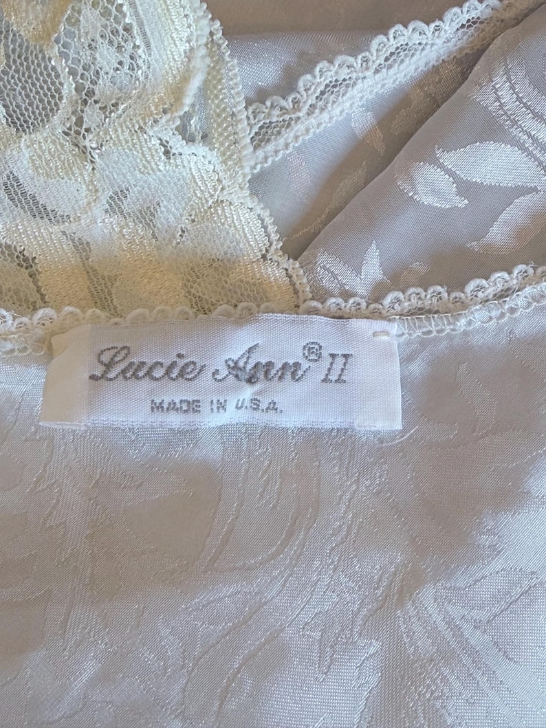 May include: A white garment featuring a tonal floral jacquard pattern and cream-colored lace trim along the neckline. A white fabric label is stitched to the garment, displaying the text 'Lucie Ann II' and 'MADE IN U.S.A.' in black lettering.