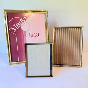 May include: Three gold-toned metal picture frames of varying sizes. The largest frame features a pink insert with the text Magee Company 8x10. The middle frame has a plain off-white insert, and the right frame contains a textured beige fabric insert.