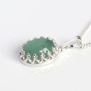 Puede incluir: Un collar con colgante de plata con una piedra preciosa verde ovalada. La piedra est&aacute; engastada en un bisel decorativo de plata en forma de corona. La cadena del collar es una fina cadena de plata.